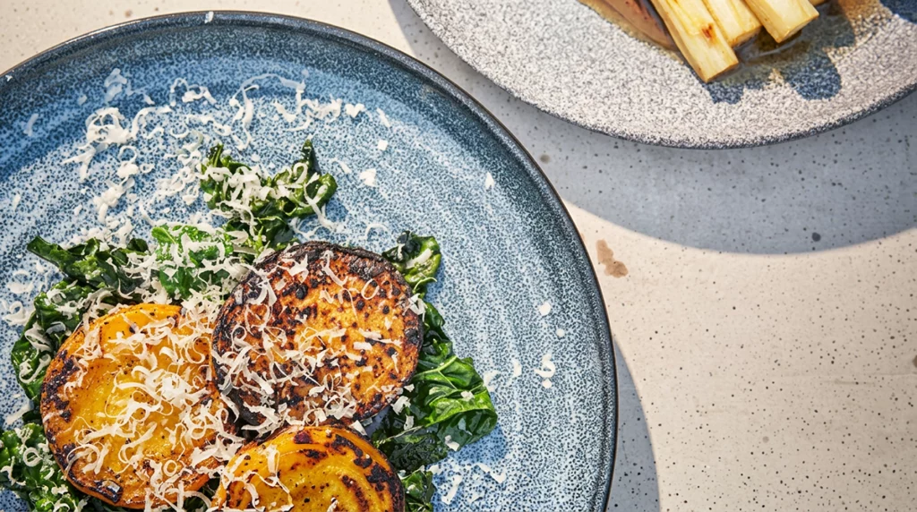 Grillade skivor av gulbeta på smörstekt svartkål, toppade med riven parmesan, på ett blått fat