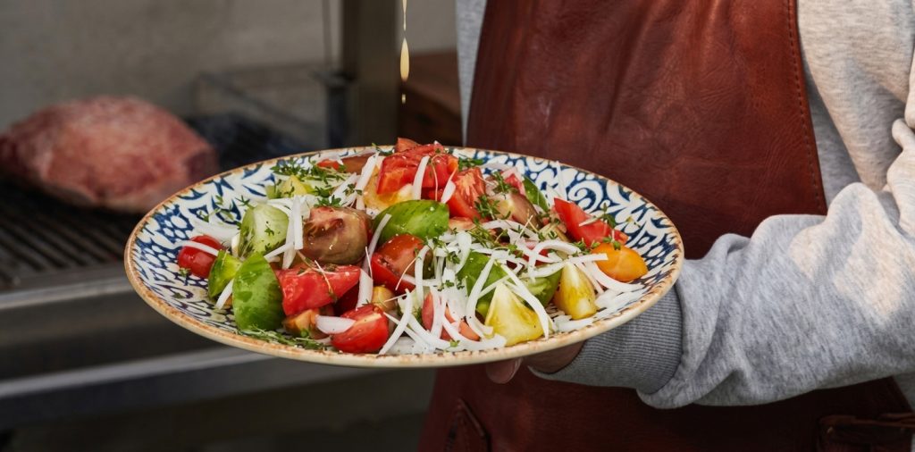 Tomatsallad med senapsvinägrett och silverlök på ett mönstrat fat, buren av en person i förkläde framför en grill