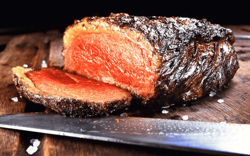 Ugnsstekt picanha med mörk stekyta, uppskuren så att det rosa köttet syns, på en skärbräda med kniv