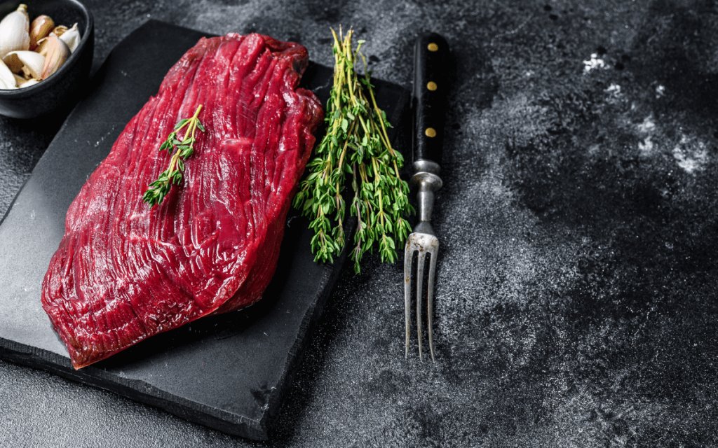 Rå bit älgkött på en skärbräda av skiffer med rosmarin, timjan och en stekgaffel