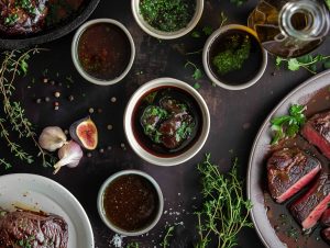 Flera såser till kött i små skålar, omgivna av skivat nötkött, fikon, vitlök och färska örter